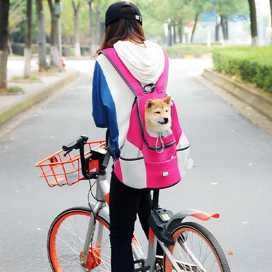 Une femme fait du vélo avec un Shiba Inu qui sort d'un sac à dos pour chien | Nidouillet ™ de transport ,rose pour , entourée d'arbres dans une rue calme.