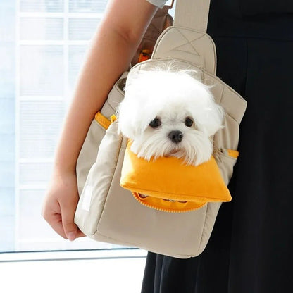 Un petit chien blanc sort d'un  sac de transport polyvalent pour chien | coupe - vente kaki avec un coussin orange, tenu par une personne en robe noire.
