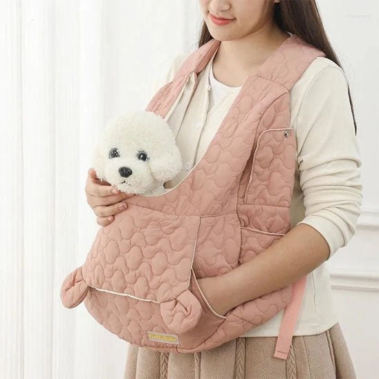Une femme tient un chien en peluche doux dans un sac de transport pour chien ventral rose, souriant sur un fond de couleur claire.